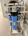 Dual Crystal Quest Big Blue SMART whole house water filtration system on a white floor stand with pressure gauge.