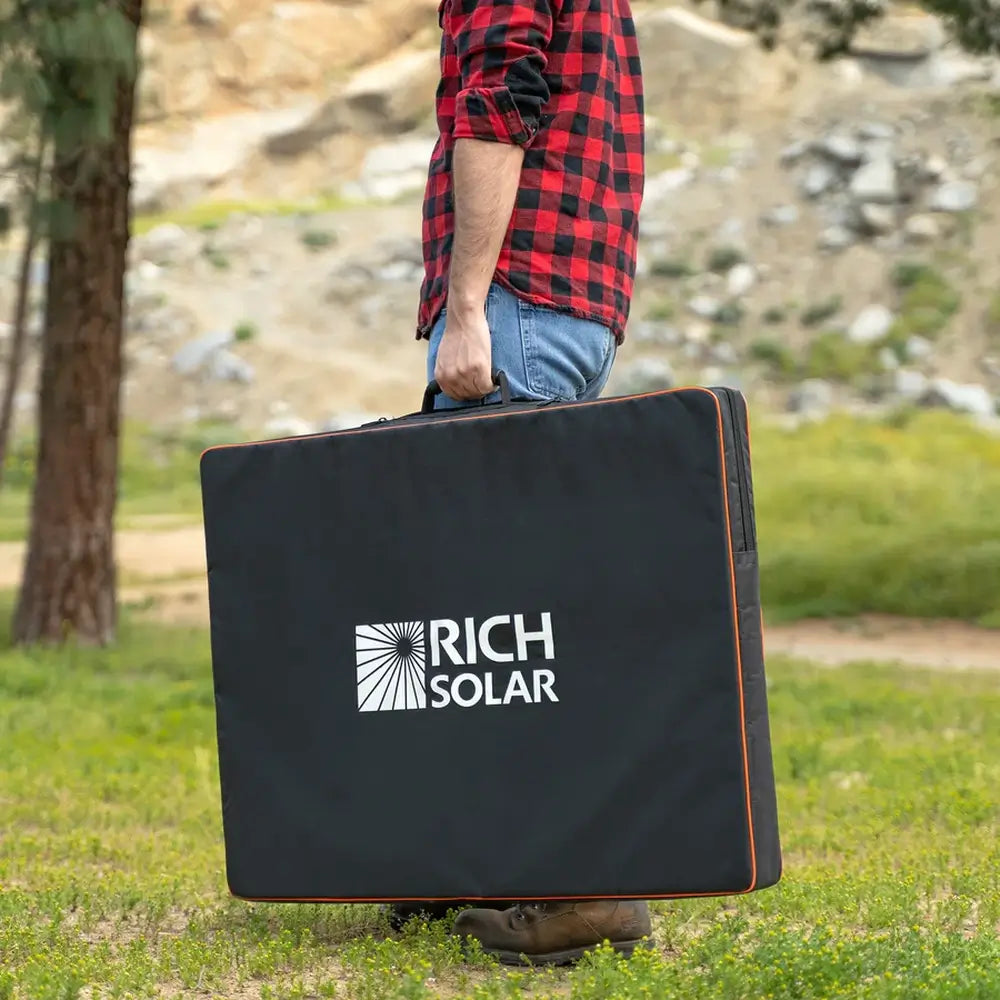 Person carrying a Rich Solar MEGA 200W portable solar panel inside its black protective zippered storage bag outdoors.