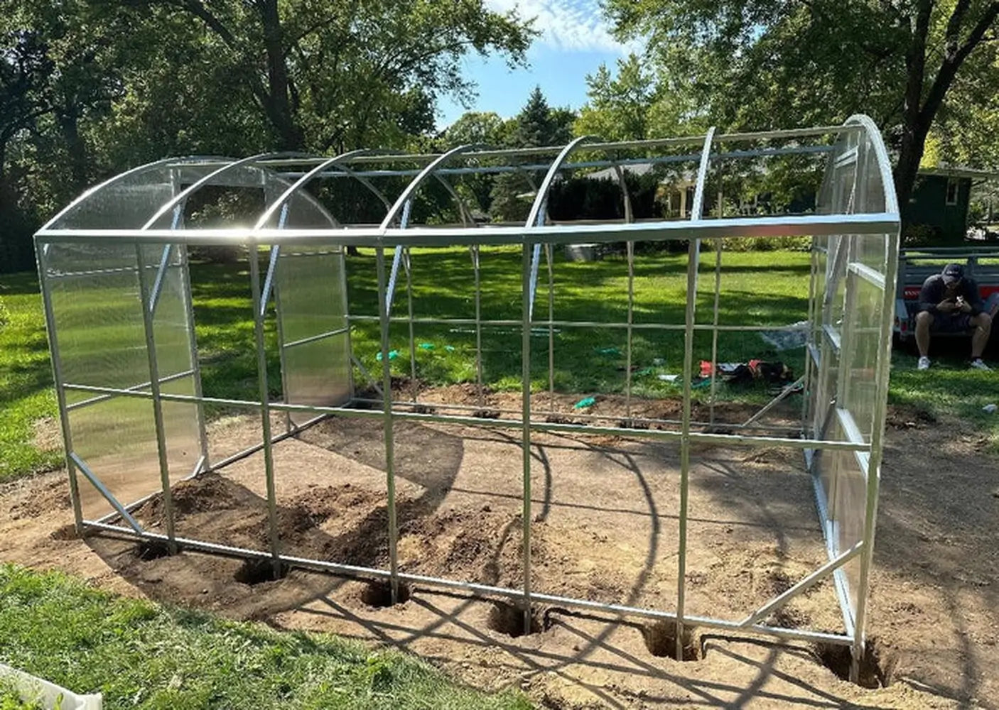 The full skeleton frame of a Magnolia 10x13 Greenhouse Kit installed in a backyard with visible ground anchor holes for stability.