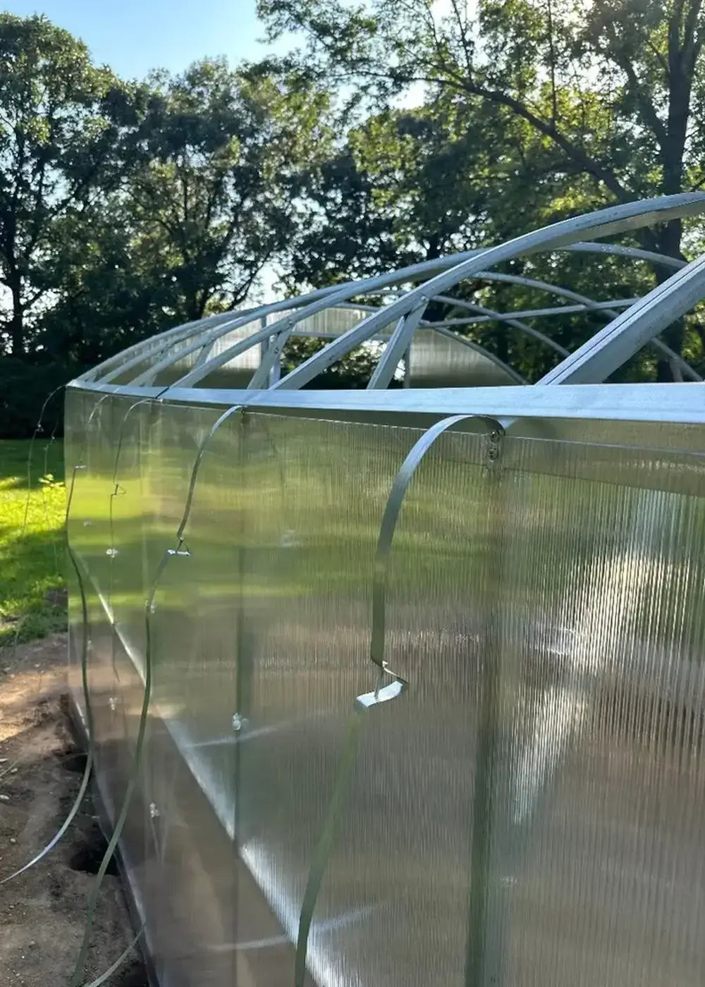 Detailed view of polycarbonate panel installation with metal securing straps on the side of a GreenhouseToGrow Magnolia greenhouse.