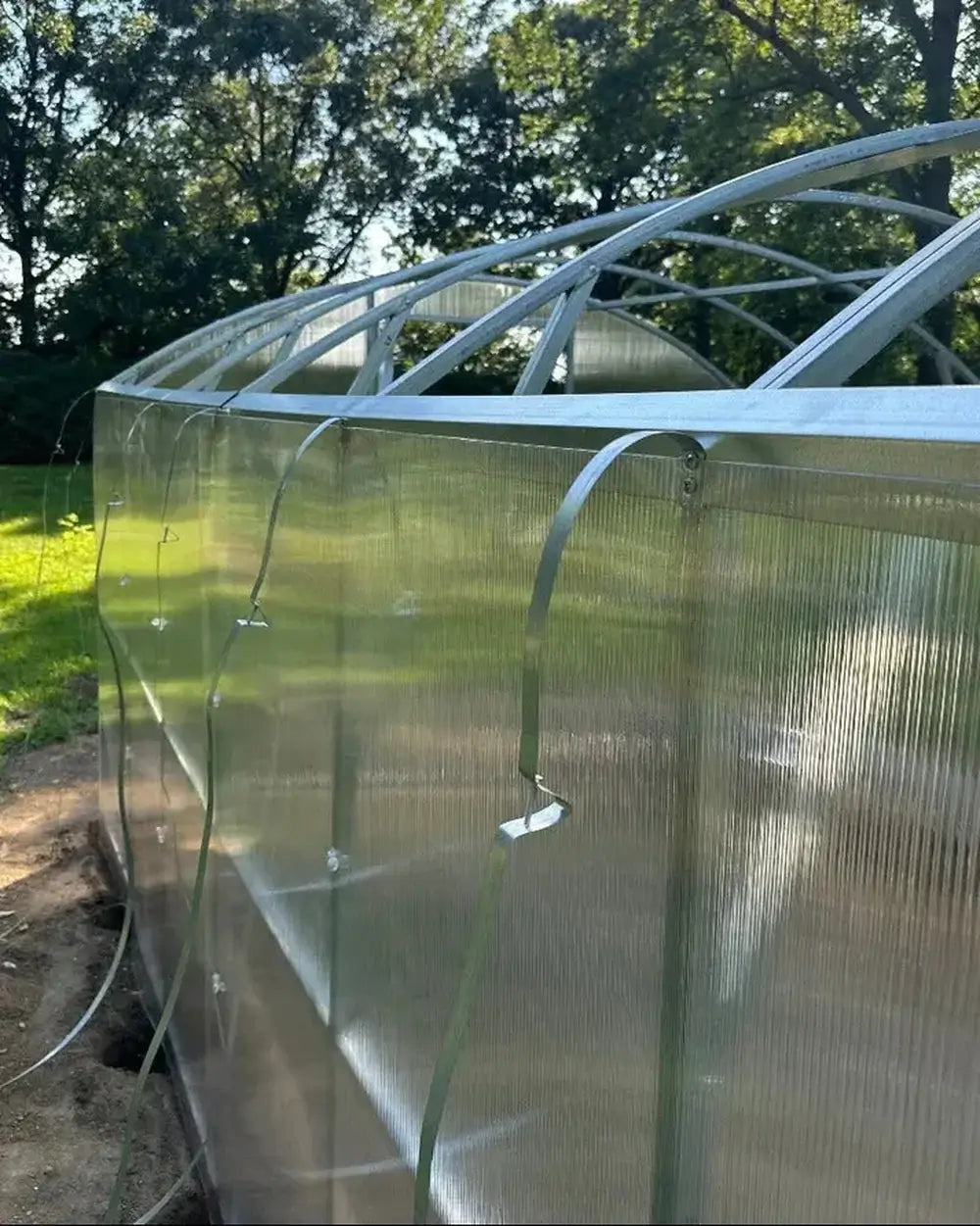 Exterior side view showing the clear polycarbonate panels of the Magnolia 10x13 Greenhouse Kit secured with metal fastening bands.