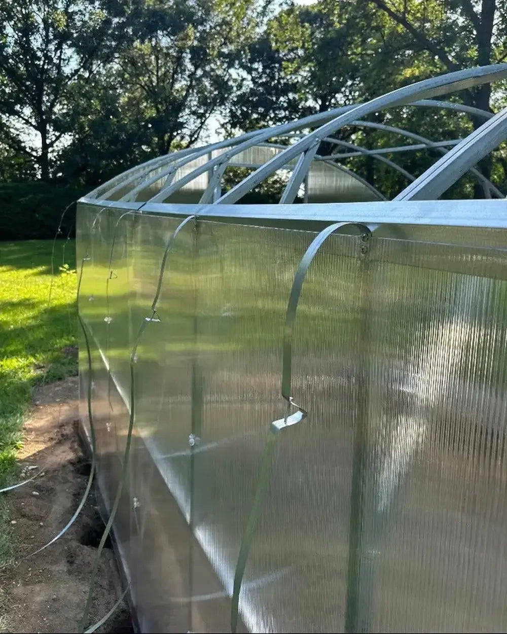 Detail of the UV-protected double-wall polycarbonate panels being secured to the 10'x26' MAGNOLIA REINFORCED greenhouse frame with metal fastening straps.