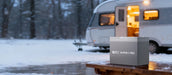 Rich Solar ALPHA 1 PRO 12V 100Ah deep cycle lithium battery resting on a snowy wooden table with a winter RV trailer in the background.