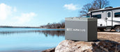 Rich Solar Alpha 1 Lite 12V 100Ah LiFePO4 battery resting on a rock by a scenic lake with an RV motorhome in the background.