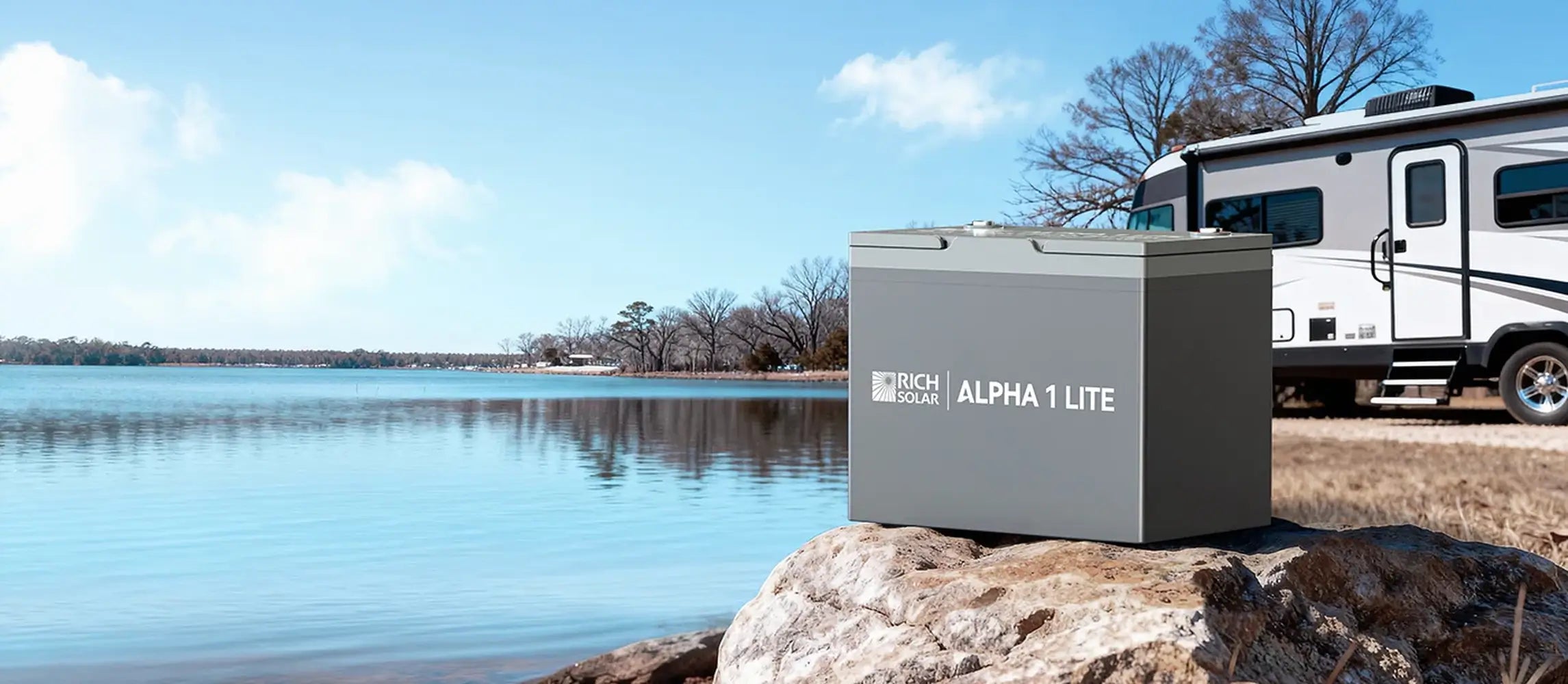 Rich Solar Alpha 1 Lite 12V 100Ah LiFePO4 battery resting on a rock by a scenic lake with an RV motorhome in the background.