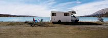 Off-grid digital nomad working on a laptop outside a white camper van RV parked next to a beautiful alpine lake.