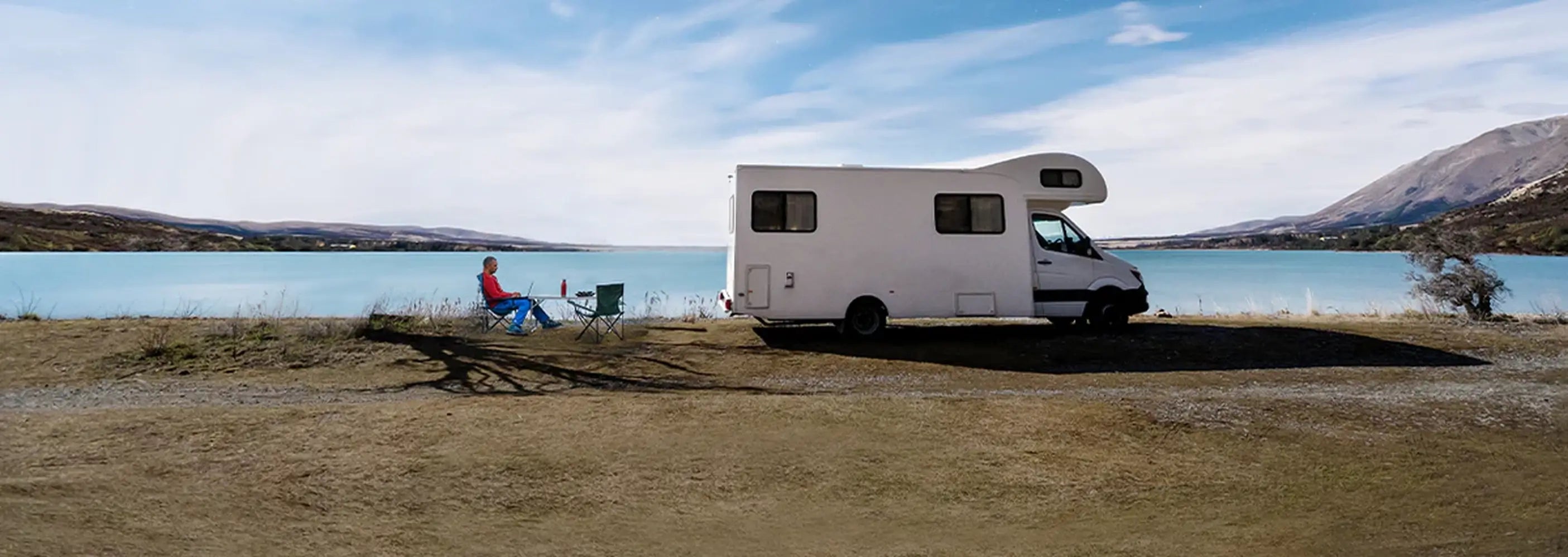 Off-grid digital nomad working on a laptop outside a white camper van RV parked next to a beautiful alpine lake.