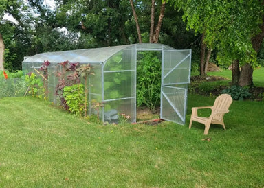 Full exterior view of an assembled GreenhouseToGrow Magnolia 10x26 Greenhouse Kit in a backyard garden with an open Dutch door and tall interior plants.