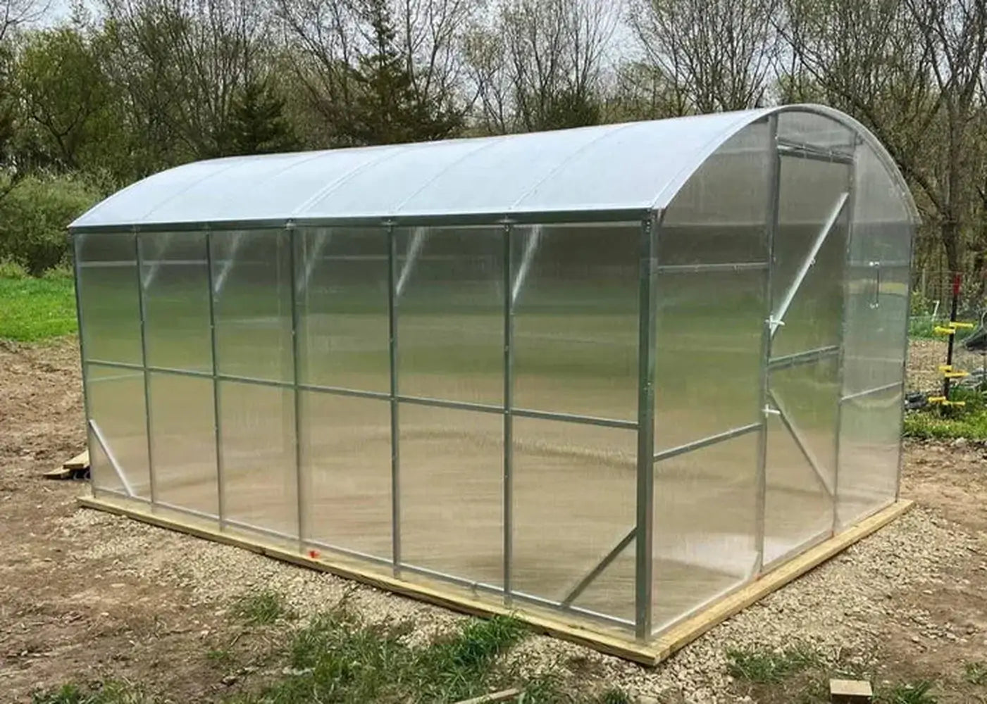 The completed GreenhouseToGrow Magnolia 10x13 Greenhouse Kit installed on a level wood and gravel foundation in a garden setting.