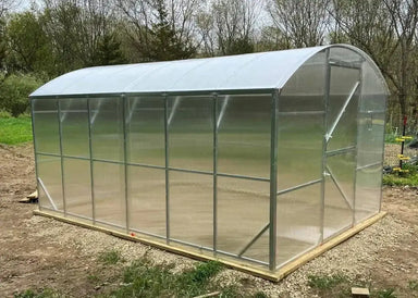 The completed GreenhouseToGrow Magnolia 10x13 Greenhouse Kit installed on a level wood and gravel foundation in a garden setting.