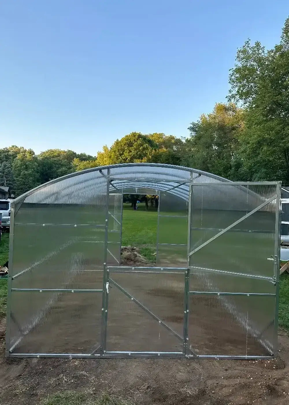 Front entrance view of a GreenhouseToGrow Magnolia 10x32.5 Greenhouse Kit with Dutch doors overlooking a grassy field.
