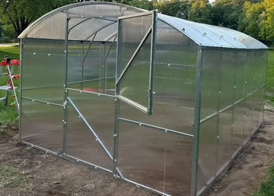 Exterior view of a fully assembled GreenhouseToGrow Magnolia 10x6.5 Greenhouse Kit with a Dutch door partially open on a backyard dirt foundation.