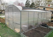 Side profile of the Magnolia 10x39 Greenhouse Kit with a protective shade cloth installed on the roof, positioned next to metal raised garden beds.
