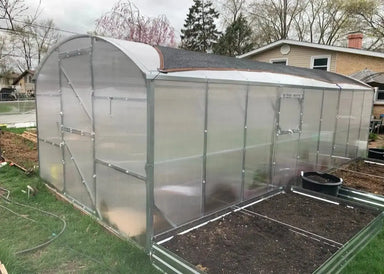 Exterior side profile of the Magnolia reinforced greenhouse 10x26 showing a side vent window and the high-density polycarbonate structure next to raised garden beds.