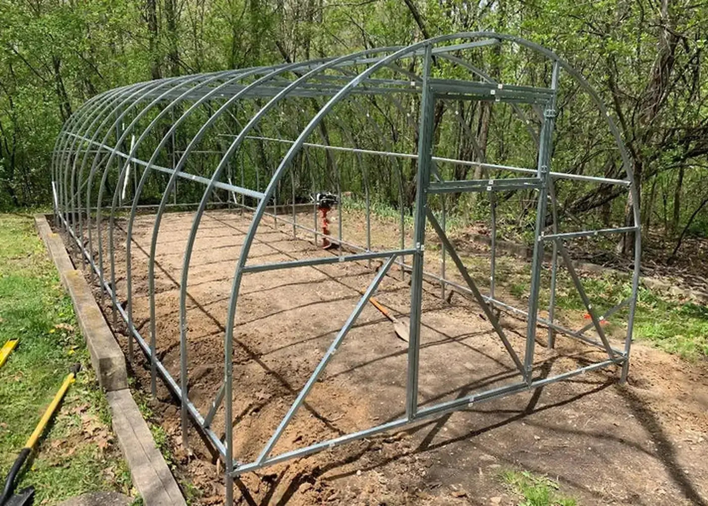 The exposed galvanized steel frame of a GreenhouseToGrow Camellia 10x13 Greenhouse Kit during the assembly process on a garden site.
