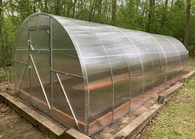 Angled exterior view of the GreenhouseToGrow Camellia Greenhouse Kit installed in a wooded backyard setting with a level timber base.