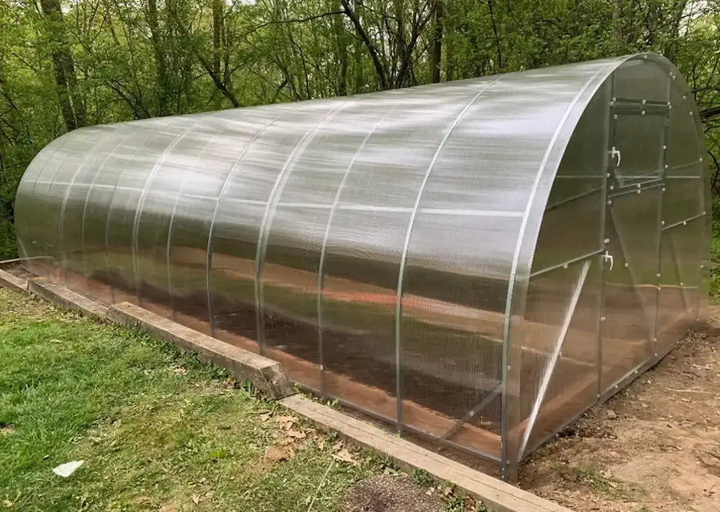Side profile of a completed Camellia 10x19.5 Greenhouse Kit installed on a timber base near a wooded area.