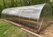 Side-angled exterior view of a completed GreenhouseToGrow Camellia 10x32.5 Greenhouse Kit, showcasing the long arched polycarbonate structure and galvanized steel frame built on a timber foundation.