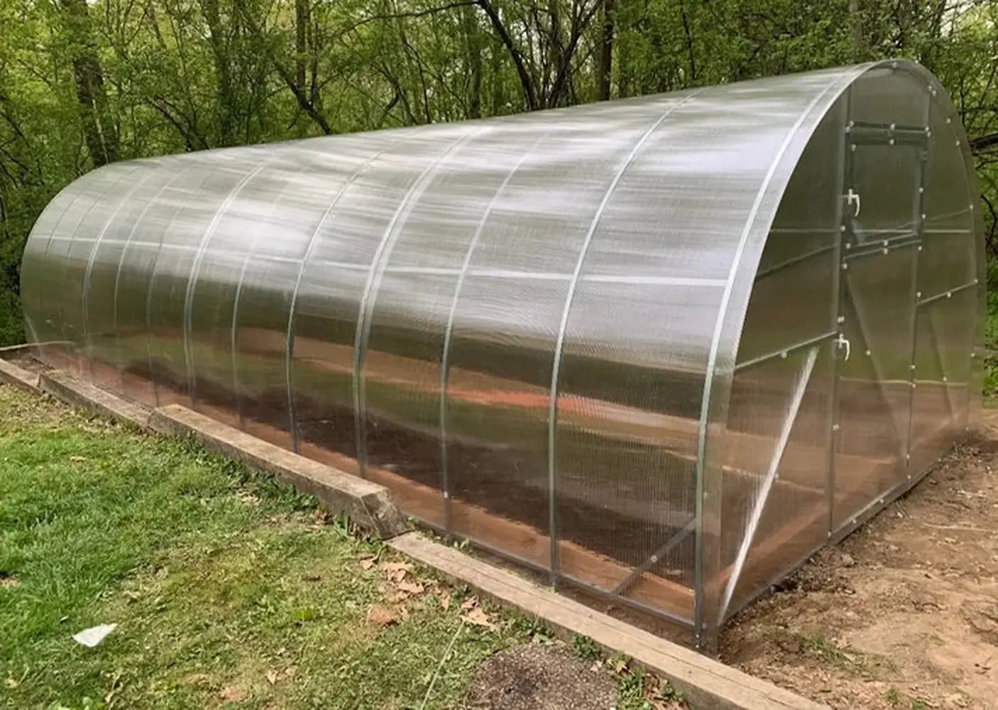 Side-angled exterior view of a completed GreenhouseToGrow Camellia 10x32.5 Greenhouse Kit, showcasing the long arched polycarbonate structure and galvanized steel frame built on a timber foundation.