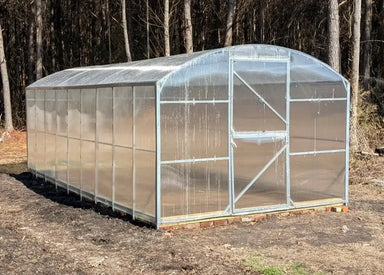 Frontal view of a finished GreenhouseToGrow Magnolia 10x32.5 Greenhouse Kit standing on a brick foundation near a wooded area.