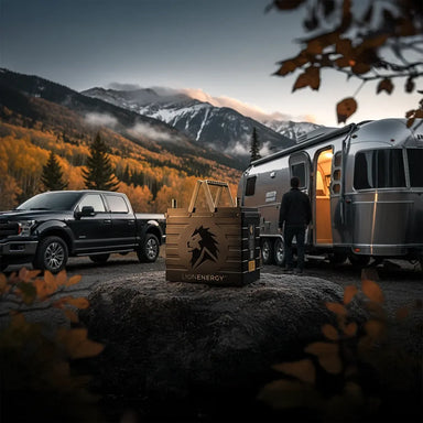Lion Energy UT 1300 BT-H battery on a rock at an RV campsite with pickup truck and silver trailer, set against misty mountains and fall foliage at dusk