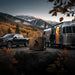 Lion Energy UT 1300 BT-H battery on a rock at an RV campsite with pickup truck and silver trailer, set against misty mountains and fall foliage at dusk