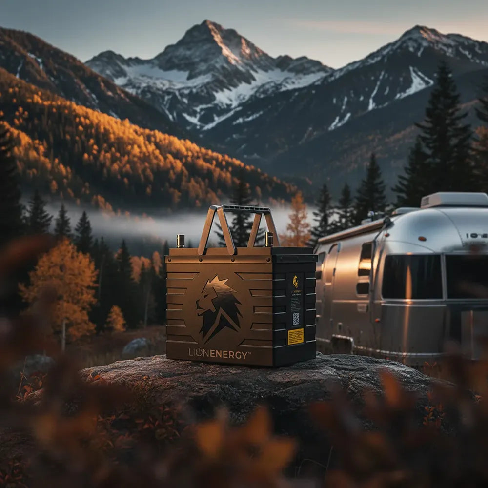 Lion Energy UT 1300 BT-H 105Ah lithium iron phosphate battery on a rock at a mountain campsite beside a silver travel trailer, with snowy peaks and autumn trees in the background
