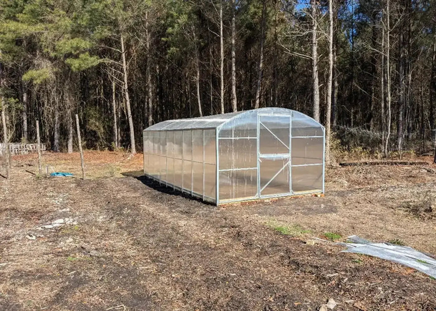 Wide-angle view of a completed GreenhouseToGrow Magnolia 10x32.5 Greenhouse Kit in a large backyard with a forest backdrop.