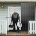 Size comparison of a massive Renogy 590W solar panel standing vertically next to a standard 80-inch residential front door, featuring a solar installer with tools to demonstrate scale.