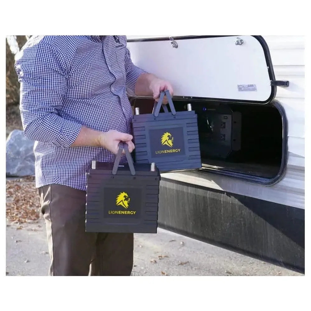 Person carrying two Lion Energy Safari UT 1300 portable batteries by their fold-up handles near an RV storage compartment, demonstrating portable off-grid power and RV battery use