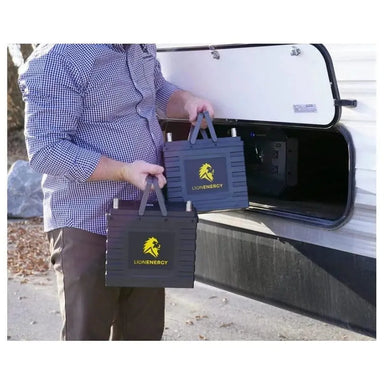 Person carrying two Lion Energy Safari UT 1300 portable batteries by their fold-up handles near an RV storage compartment, demonstrating portable off-grid power and RV battery use